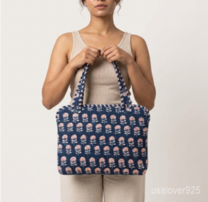 Close-up of the handcrafted blue floral block print detailing, dense quilted stitching texture lines, robust handle strap attachments, and clean zip track on an Indian cotton MacBook case.